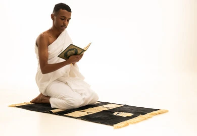 Memorizing and reciting the Holy Quran, feeling tranquility and comfort, a portrait of a young Saudi Arabian Gulf man wearing Ihram clothing sitting on a prayer rug holding the Holy Quran in his hand, performing the rituals of Hajj and Umrah, white background.