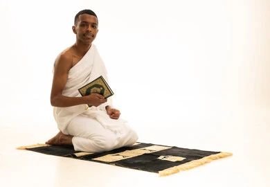 ايماءات الخشوع والطمأنينة، الشعور بالسكينة والراحة، بورتريه لشاب عربي خليجي سعودى يرتدي ثياب الإحرام يجلس على سجادة الصلاة يحمل بيده المصحف الشريف، أداء مناسك الحج والعمرة، خلفية بيضاء