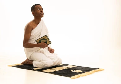 The concept of worship and drawing closer to God, the feeling of tranquility and comfort, a portrait of a young Saudi Arabian Gulf man wearing Ihram clothing sitting on a prayer rug holding the Holy Quran in his hand, looking up, performing the rituals of Hajj and Umrah, white background.