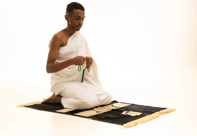 بورتريه لشاب عربي خليجي سعودى يرتدي ثياب الإحرام يجلس على سجادة الصلاة يحمل بيده السبحة الخضراء،  التسبيح والتهليل وذكرالله، أداء مناسك الحج والعمرة، خلفية بيضاء