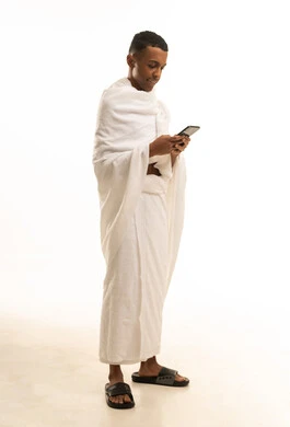Using the electronic guide application for pilgrims on a mobile phone, a portrait of a young Saudi Arabian Gulf man wearing Ihram clothing using the phone, the fifth pillar of Islam, full-body image, white background.