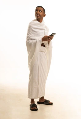 Looking at something, using the electronic guide application for pilgrims on a mobile phone, a portrait of a young Saudi Arabian Gulf man wearing Ihram clothing using the phone, the fifth pillar of Islam, full-body image, white background.