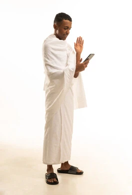 A portrait of a young Saudi Arabian Gulf man wearing Ihram clothing making a video call via mobile, visiting the Sacred House of God in Mecca, the fifth pillar of Islam, full-body image, white background.