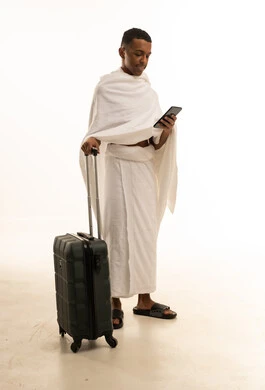 A portrait of a young Saudi Arabian Gulf man wearing Ihram clothing, holding a travel bag in one hand and browsing his phone, preparing to head out and embark on a visit to the Sacred House of God, going to perform the pilgrimage of Hajj and Umrah, full-body shot, white background.