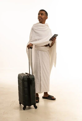 الاستعداد للتوجه والانطلاق لزيارة بيت الله الحرام، الذهاب لأداء فريضة الحج والعمرة، بورتريه لشاب عربي خليجي سعودى يرتدي ثياب الإحرام ويمسك بيده حقيبة السفر و يحمل بيده الهاتف، النظر الى شىء ما، الجسم بالطول الكامل، خلفية بيضاء