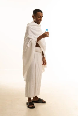 Increasing fluid intake to keep the body hydrated, performing the rituals of Hajj and Umrah, a portrait of a young Saudi Arabian Gulf man wearing Ihram clothing holding a water bottle in his hand, full-body image, white background.