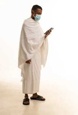 بورتريه من الجانب لشاب عربي خليجي سعودى يرتدي ثياب الإحرام والكمامة يتصفح الهاتف المحمول، الدليل الالكتروني للحاج والمعتمر، الوقاية من فايروس كورونا، صورة الجسم بالطول الكامل، خلفية بيضاء