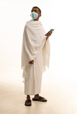 الدليل الالكتروني للحاج و المعتمر، بورتريه لشاب عربي خليجي سعودى يرتدي ثياب الإحرام والكمامة يستخدم الهاتف المحمول، الوقاية من فايروس كورونا، النظر اشىء ما، صورة الجسم بالطول الكامل، خلفية بيضاء