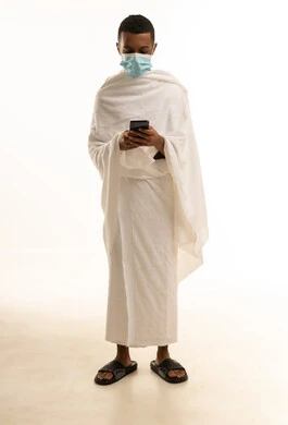 استخدام تطبيق دليل الحاج والمعتمرالالكتروني على الهاتف المحمول، بورتريه لشاب عربي خليجي سعودى يرتدي ثياب الإحرام والكمامة يحمل بيده الجوال، الوقاية من فايروس كورونا، صورة الجسم بالطول الكامل، خلفية بيضاء