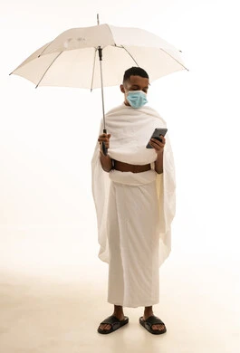 التقرب الى الله بأداء الطاعات، الوقاية من فايروس كورونا، آداء مناسك الحج والعمرة، بورتريه لشاب عربي خليجي سعودى يرتدي ثياب الإحرام والكمامة يحمل بيده الهاتف المحمول ومظلة بيضاء، صورة الجسم بالطول الكامل، خلفية بيضاء