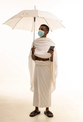 الوقاية من فايروس كورونا، آداء مناسك الحج والعمرة، بورتريه لشاب عربي خليجي سعودى يرتدي ثياب الإحرام والكمامة يحمل بيده الهاتف المحمول ومظلة بيضاء، صورة الجسم بالطول الكامل، خلفية بيضاء