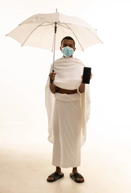 Performing the rituals of Hajj and Umrah, a portrait of a young Saudi Arabian Gulf Arab man wearing Ihram clothing and a mask, holding a mobile phone with a blank black screen and a white umbrella, for protection against the coronavirus, full-body image, white background.