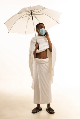 Social media and social relations, a portrait of a young Arab Gulf Saudi man wearing Ihram clothing and a mask, making a phone call and holding a white umbrella, performing the rituals of Hajj and Umrah, prevention from the Coronavirus, white background.
