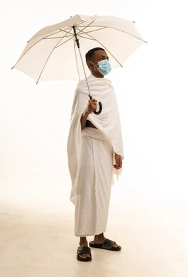 الركن الخامس من أركان الإسلام، بورتريه لشاب عربي خليجي سعودى يرتدي ثياب الإحرام و الكمامة يحمل بيده مظلة حامية من المطر، آداء مناسك الحج والعمرة، الوقاية من فايروس كورونا، خلفية بيضاء
