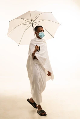 آداء مناسك الحج والعمرة، بورتريه من الجانب لشاب عربي خليجي سعودى يرتدي ثياب الإحرام و الكمامة يحمل بيده مظلة حامية من المطر،  الوقاية من فايروس كورونا, الركن الخامس من أركان الاسلام، خلفية بيضاء