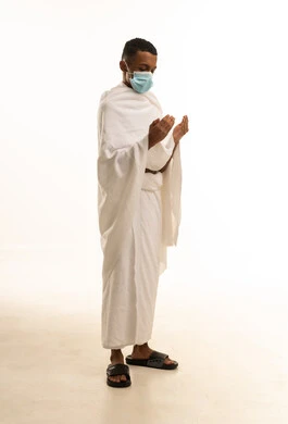 موسم العمرة في شهر رمضان المبارك، الدعاء والعبادة والتقرب إلى الله، بورتريه من الجانب لشاب عربي خليجي سعودى يرتدي ثياب الإحرام والكمامة يرفع يديه للأعلى يدعو بتضرع وخشوع، إجراءات الوقاية من فايروس كورونا، خلفية بيضاء