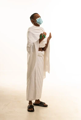 الركن الخامس من أركان الإسلام، بورتريه من الجانب لشاب عربي خليجي سعودى يرتدي ثياب الإحرام والكمامة يرفع يديه للأعلى يدعو بتضرع وخشوع، الدعاء والتقرب إلى الله، أداء مناسك الحج والعمرة، إجراءات الوقاية من فايروس كورونا، خلفية بيضاء