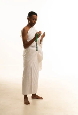 Supplication and drawing closer to God, a side portrait of a young Saudi Arabian Gulf man wearing Ihram clothing and holding a prayer beads, raising his hands upwards in supplication with humility and reverence, the concept of the pillars of Islam, performing the rituals of Hajj and Umrah, white background.