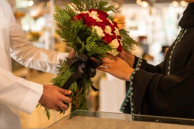 الزوج يقدم لزوجته باقة ورد , زوجان عربيان خليجيان سعوديان سعيدان يحتفلان بعيد الحب ,الزوجة تعبر بإيماءات وجه ويدين تدل على الدهشة والفرح , يوم عيد الفلانتاين , أعياد ومناسبات سعيده , الاحتفال بعيد الميلاد , خلفية محل بيع الورود.