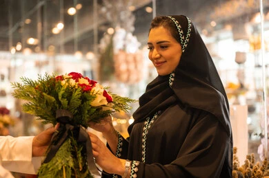 الاحتفال بعيد الميلاد , زوجان عربيان خليجيان سعوديان سعيدان يحتفلان بعيد الحب , الزوج يقدم لزوجته باقة ورد , الزوجة تعبر بإيماءات وجه ويدين تدل على الدهشة والفرح , يوم عيد الفلانتاين , أعياد ومناسبات سعيده , خلفية محل بيع الورود.