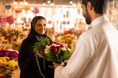 أعياد ومناسبات سعيده , زوجان عربيان خليجيان سعوديان سعيدان يحتفلان بعيد الحب , الزوج يقدم لزوجته باقة ورد , الزوجة تعبر بإيماءات وجه ويدين تدل على الدهشة والفرح , يوم عيد الفلانتاين ,الاحتفال بعيد الميلاد , خلفية محل بيع الورود.