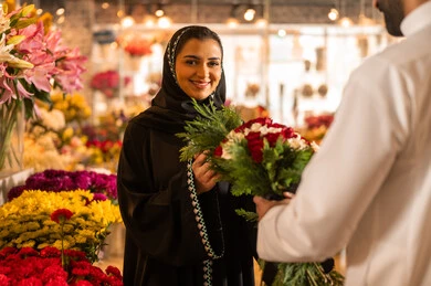 الاحتفال بعيد الميلاد , زوجان عربيان خليجيان سعوديان سعيدان يحتفلان بعيد الحب , الزوج يقدم لزوجته باقة ورد , الزوجة تعبر بإيماءات وجه ويدين تدل على الدهشة والفرح , يوم عيد الفلانتاين , أعياد ومناسبات سعيده , خلفية محل بيع الورود.