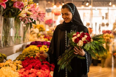 امرأة عربية خليجية سعودية ترتدي العباءة والحجاب تحمل باقة ورد احمر طبيعي بايماءات الفرح والسعاده، شراء الورود والزهور الطبيعية للأعياد والمناسبات من المحلات والمتاجر