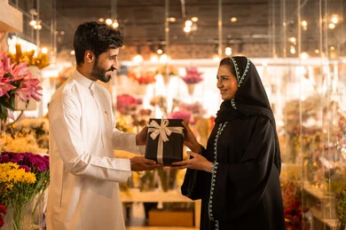 Celebrating Christmas, the concept of affection and intimacy, a happy Saudi Arabian Gulf couple celebrating Valentine's Day, the husband presents his wife with a gift box, the wife expresses with facial and hand gestures indicating surprise and joy, Valentine's Day, happy holidays and occasions, background of a flower shop.