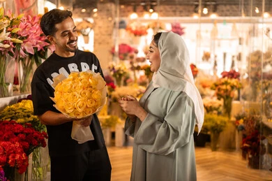 The husband presents his wife with a bouquet of flowers, a happy Saudi Arabian couple celebrating Valentine's Day, the wife expresses surprise and joy through facial and hand gestures, Valentine's Day, happy holidays and occasions, celebrating Christmas, background of a flower shop.