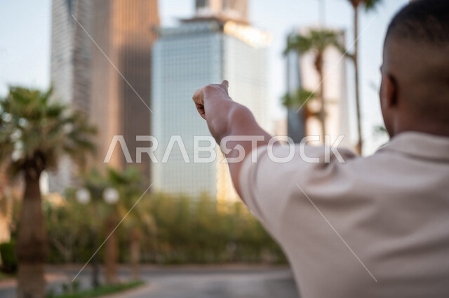 صورة من الخلف لشاب عربي خليجي سعودي أسمر يقف و يشير إلى أبراج مدينة الملك عبدالله المالية ( كافد ) في مدينة الرياض في المملكة العربية السعوية  ، إطلالة رائعة لأبراج المدينة ، جولة سياحية في المدينة