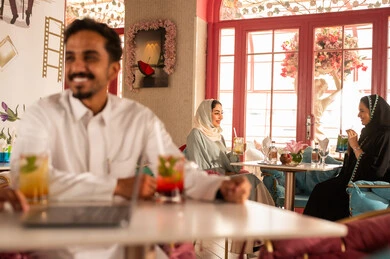 Completing work tasks abroad, spending enjoyable times with friends, discussing and conversing in calm atmospheres, sharing cold drinks, two Arab Gulf friends from Saudi Arabia wearing white thobes are sitting in one of the upscale cafes using a laptop, utilizing modern technology.