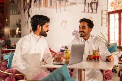 Completing work tasks abroad, spending enjoyable times with friends, discussing and conversing in calm atmospheres, sharing cold drinks, two Arab Gulf friends from Saudi Arabia wearing white thobes are sitting in one of the upscale cafes using a laptop, utilizing modern technology.