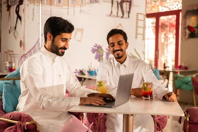 انجاز مهام العمل في الخارج، قضاء أوقات ممتعة مع الأصدقاء، النقاش والحوار في الأجواء الهادئة، مشاركة المشروبات الباردة ، صديقان عربيان خليجيان سعوديان يرتديان الثوب الابيض يجلسان  في إحدى المقاهي الراقية يقومان باستخدام الحاسوب المحمول، استخدام جهاز تقني حديث