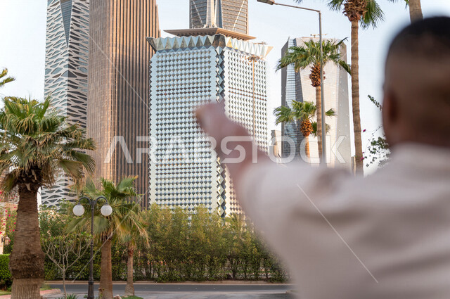 صورة من الخلف لشاب عربي خليجي سعودي يقف و يشير إلى أبراج مدينة الملك عبدالله المالية ( كافد ) في مدينة الرياض في المملكة العربية السعوية  ، إطلالة رائعة لأبراج المدينة ، جولة سياحية في المدينة