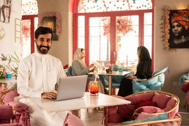 A Saudi Arabian Gulf man wearing a white thobe is sitting in a high-end café using a laptop, working remotely, completing tasks, managing and organizing business affairs through the laptop, enjoying cold drinks, and using a modern and advanced technological device.