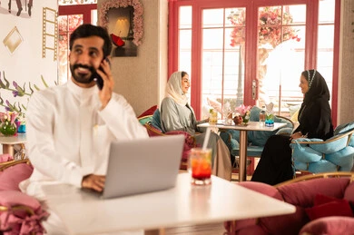 Making a phone call, working remotely, completing tasks and managing and organizing business affairs via a laptop, enjoying cold drinks, using a modern and advanced technological device, an Arab Gulf Saudi man wearing a white thobe sitting in a luxury café using a laptop.