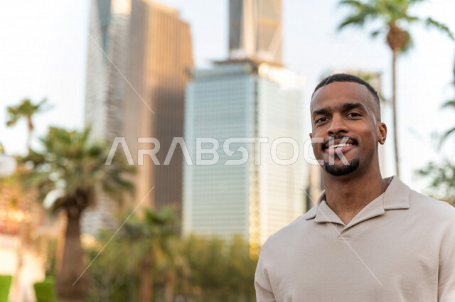 شاب عربي خليجي سعودي ذو بشرة سمراء مبتسم يقف أمام أبراج مدينة الملك عبدالله المالية ( كافد ) في مدينة الرياض في المملكة العربية السعوية  ، إطلالة رائعة لأبراج المدينة ، جولة سياحية في المدينة