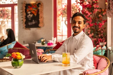 A Saudi Arabian Gulf man wearing a white thobe is sitting in a high-end café using a laptop, working remotely, completing tasks, managing and organizing business affairs through the laptop, enjoying cold drinks, and using a modern and advanced technological device.
