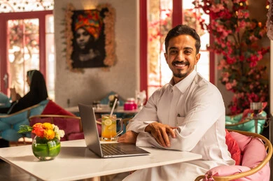 Remote work, completing tasks and managing business affairs and organizing them via laptop, enjoying cold drinks, using a modern and advanced technical device, an Arab Gulf Saudi man wearing a white thobe sitting in one of the upscale cafes and using a laptop.