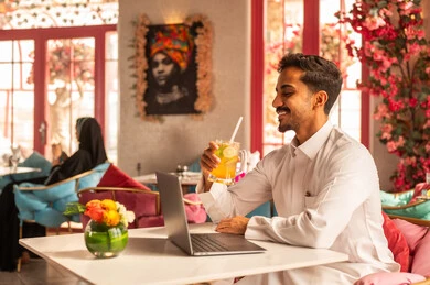 Using a modern and advanced technical device, working remotely, completing tasks and managing business affairs and organizing them via a laptop, enjoying cold drinks, an Arab Gulf Saudi man wearing a white thobe sitting in one of the upscale cafes using a laptop.