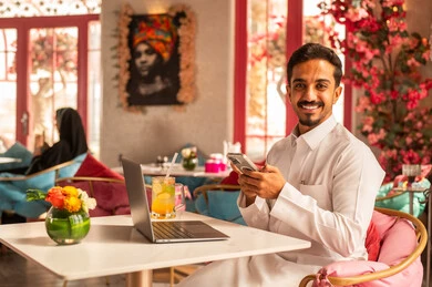 Making a phone call, working remotely, completing tasks and managing and organizing business affairs via a laptop, enjoying cold drinks, using a modern and advanced technological device, an Arab Gulf Saudi man wearing a white thobe sitting in a luxury café using a laptop.