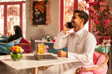 اجراء مكالمة هاتفية عبر الهاتف، العمل عن بعد، انجاز المهام وإدارة شؤون الأعمال وتنظيمها عبر اللابتوب، الاستمتاع بشرب المشروبات الباردة ، استخدام  جهاز تقني حديث ومتطور، رجل عربي خليجي سعودي يرتدي الثوب الابيض يجلس فى احدي المقاهي الراقيه  ويستخدم الحاسوب المحمول