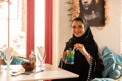 Spending enjoyable times with friends, an upscale coffee shop in the Kingdom of Saudi Arabia, expressions of happiness and joy, discussion and dialogue in a calm atmosphere, sharing cold drinks, two Arab Gulf Saudi friends wearing abayas and hijabs sitting in one of the upscale cafes.