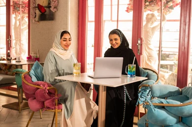 استخدام جهاز تقني حديث ، انجاز مهام العمل في الخارج، قضاء أوقات ممتعة مع الأصدقاء، النقاش والحوار في الأجواء الهادئة، مشاركة المشروبات الباردة ، صديقتان عربيتان خليجيتان سعوديتان ترتديان العباءة والحجاب تجلسان  في إحدى المقاهي الراقية تقومان باستخدام الحاسوب المحمول