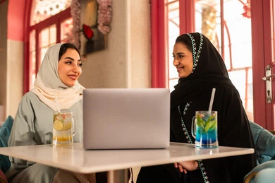 النقاش والحوار في الأجواء الهادئة، انجاز مهام العمل في الخارج، قضاء أوقات ممتعة مع الأصدقاء،مشاركة المشروبات الباردة ، صديقتان عربيتان خليجيتان سعوديتان ترتديان العباءة والحجاب تجلسان  في إحدى المقاهي الراقية تقومان باستخدام الحاسوب المحمول، استخدام جهاز تقني حديث