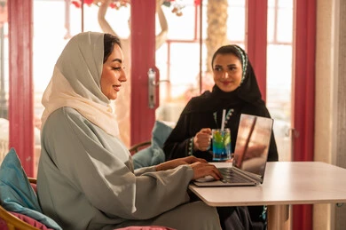 انجاز مهام العمل في الخارج، قضاء أوقات ممتعة مع الأصدقاء، النقاش والحوار في الأجواء الهادئة، مشاركة المشروبات الباردة ، صديقتان عربيتان خليجيتان سعوديتان ترتديان العباءة والحجاب تجلسان  في إحدى المقاهي الراقية تقومان باستخدام الحاسوب المحمول، استخدام جهاز تقني حديث