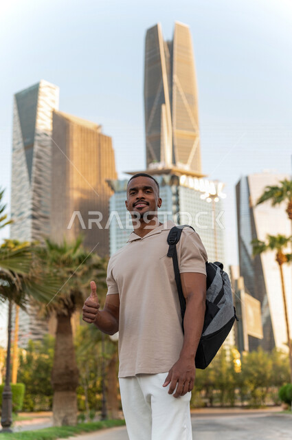 شاب عربي خليجي سعودي أسمر مبتسم يرتدي حقيبة ، يشير بإصبع الإبهام لأعلى للإعجاب  والرضى للعمل الممتاز يقف أمام أبراج مدينة الملك عبدالله المالية ( كافد ) في مدينة الرياض في المملكة العربية السعوية  ، إطلالة رائعة لأبراج المدينة ، جولة سياحية