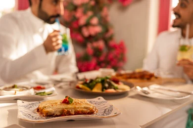 Expressions of joy and happiness, exchanging conversations and news, a lunch invitation for two Arab Gulf friends from Saudi Arabia who are enjoying food at a luxury restaurant, a dining table filled with a variety of delicious dishes and appetizers, gestures of happiness spending enjoyable times with friends, a gathering of friends at a delicious breakfast table.