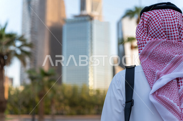 صورة من الخلف لشاب عربي خليجي سعودي يرتدي الزي التقليدي يقف أمام أبراج مدينة الملك عبدالله المالية ( كافد ) في مدينة الرياض في المملكة العربية السعودية ، إطلالة رائعة لأبراج المدينة ، جولة سياحية في المدينة