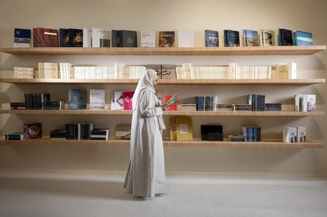 الاستمتاع بقراءة الكتب والقصص والروايات, ثقافة التعلم الذاتي, إنجاز الواجبات اليومية, قضاء الوقت بالقراءة والتعلم والتثقف، امرأة عربية سعودية خليجية محجبة ترتدي العباءة تحمل بيديها كتاب تقف داخل معرض الكتاب، ارفف مليئة بالكتب المختلفة